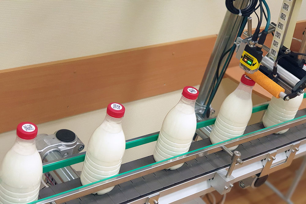 Верхнее нанесение стикеров с кодом дата матрикс на цилиндрические молочные бутылочки движуюсия на конвейере