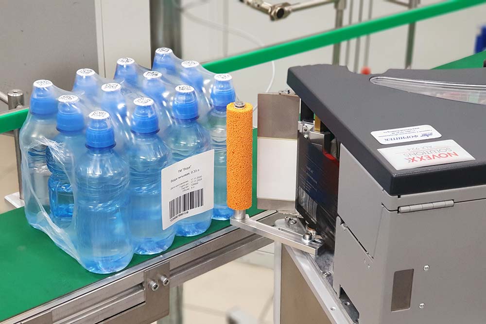 Нанесение этикетки сбоку принтером-аппликатором на упаковку воды на конвейере